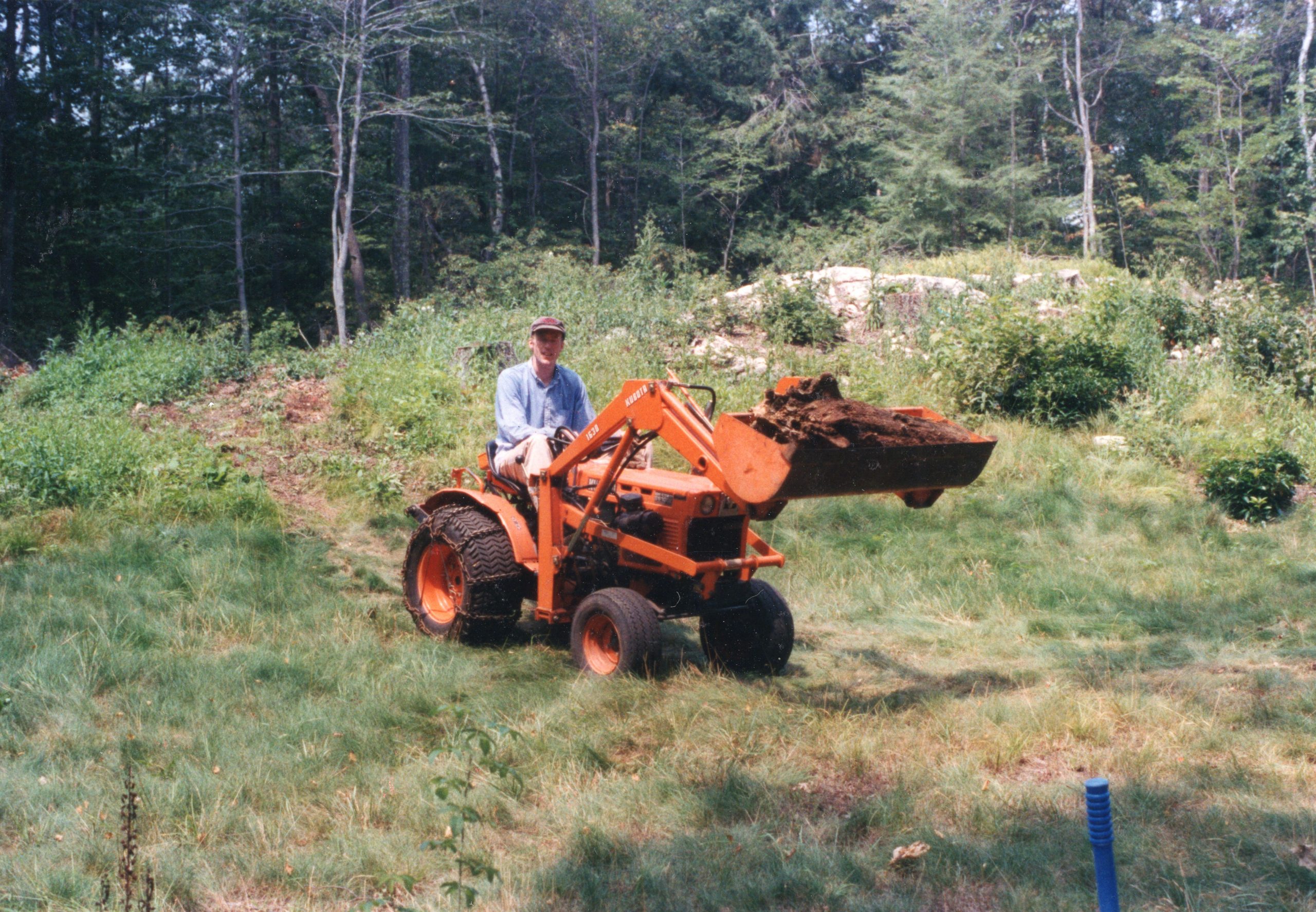 Man driving an orange Kubota tractor with a bucket loader. ROPS not yet installed. Rock in background matches description in article.